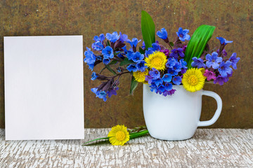 Bouquet of primroses blue lungwort in white enameled metal mug on table and vintage background, grunge with white paper sheet for text, vintage, copy space, mock up
