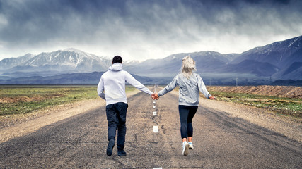 blonde girl with a guy running on the road holding hands on the background of black snow-covered mountains in the clouds