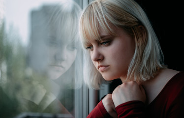 Portrait of depressed young blond woman near window at home. Sadness, nostlagic, depression. - obrazy, fototapety, plakaty