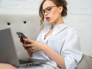 Portrait of a serious young woman holding mobile phone while sit in bed at home with laptop computer.