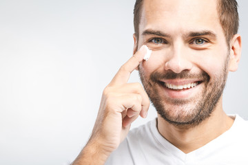 Skin care. Handsome young shirtless man applying cream at his face and looking at himself with smile while standing over gray background and looking at camera. Close-Up. Space for text.