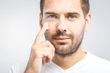 Skin care. Handsome young shirtless man applying cream at his face and looking at himself with smile while standing over gray background and looking at camera. Close-Up. Space for text.