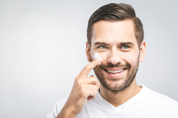 Skin care. Handsome young shirtless man applying cream at his face and looking at himself with smile while standing over gray background. Close-Up. Space for text.