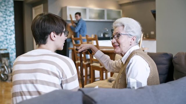 PAN of cheerful young woman with short hair sitting on sofa and chatting with elderly woman with walking stick; unrecognizable man unpacking groceries in background