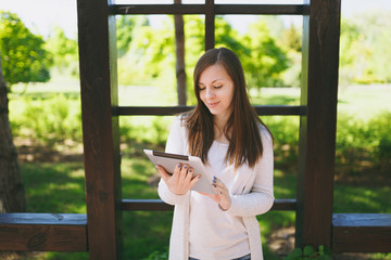Fototapeta premium Portrait of beautiful woman with charming smile wearing light casual clothes. Pretty girl holding tablet pc computer, reading news in city park in street outdoors on spring nature. Lifestyle concept.