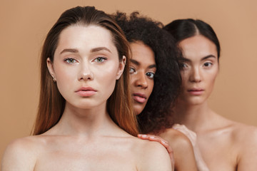 Beauty portrait of three nude women of different nation: caucasian, african american and asian girls, standing together isolated over beige background