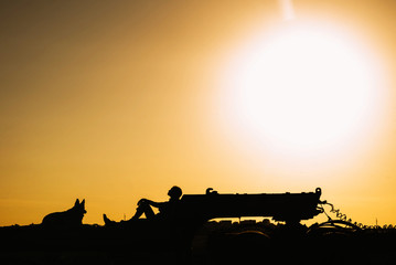Fototapeta premium The girl is sitting on a huge trailer at sunset, and her dog is next door