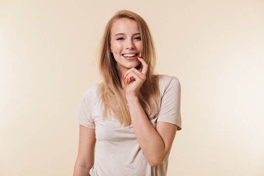 Photo of elegant woman 20s with european appearance in basic t-shirt smiling at camera and holding finger at face, isolated over beige background in studio