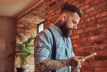 Fototapeta premium Old-fashioned tattooed hipster wearing a shirt and suspenders, in a sunglasses, using a tablet while standing near a desk in an office with loft interior.