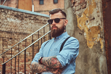 Stylish handsome old-fashioned tattooed hipster guy in a shirt with suspenders, standing near an...