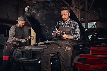 Two bearded auto mechanic in a uniform, repair a broken car in the garage. 