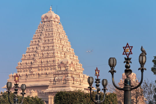 View of lamps with a traditional Hindu six-pointed star, and the main building of the Chattarpura temple complex near by which the passenger plane flies by.