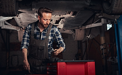 Fototapeta na wymiar Auto mechanic in a uniform, working on a workbench while standing under lifting car in a repair garage. 