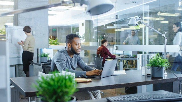 Busy International Office, African-American Man Working at His Desk on a Laptop, He Writes in Reminder His Notebook. In Background Creative Young People Working.
