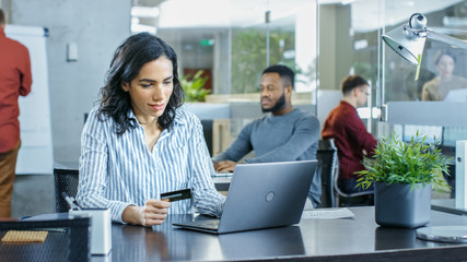 Beautiful Female Office Worker Makes Internet Purchase from Her Laptop Using Credit Card. In the Background Creative Young People Working.
