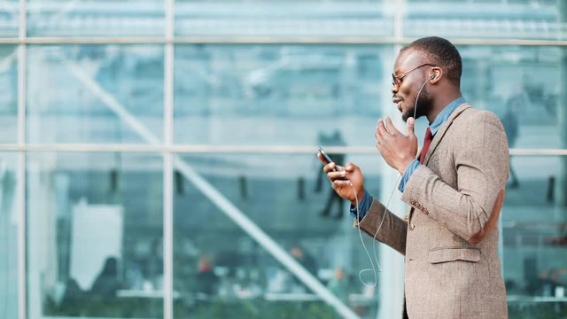 African American businessman in the sunglasses talking on the phone with headphones near the office building. Business, people, music, technology, leisure and lifestyle concept