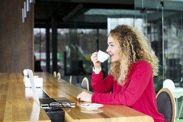 Beautiful blonde young woman drinking cappuccino coffee in a hipster bar and smiling. Curly haired female blogger, smiling, surfing, browsing, blogging on laptop computer. Windows background, portrait
