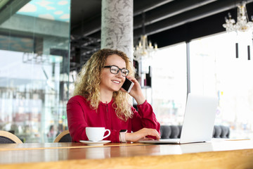 Fototapeta na wymiar Attractive young woman sitting at coffee shop with her fashionable laptop, drinking cappuccino, talking on the phone, receiving good news smiling. Beautiful blonde female with curly hair. Background.