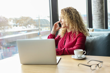 Attractive young woman sitting at coffee shop with her fashionable laptop, drinking cappuccino, talking on the phone, receiving good news smiling. Beautiful blonde female with curly hair. Background.