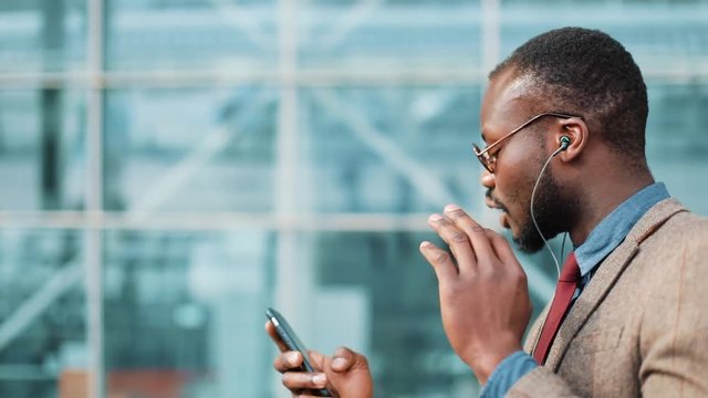 African American businessman in the sunglasses talking on the phone with headphones near the office building. Business, people, music, technology, leisure and lifestyle concept