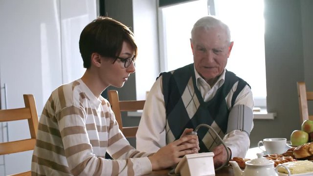 Zoom out shot of young woman in glasses talking to senior man sitting at kitchen table and measuring his blood pressure using sphygmomanometer