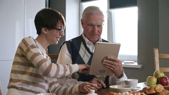 PAN of laughing young woman in glasses helping her senior grandfather with tablet while having breakfast in morning: she showing him how to use it and explaining something