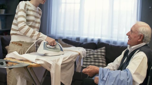 Tilt up of cheerful young woman in glasses ironing shirts and chatting with senior man in wheelchair