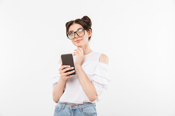 Photo of dreaming woman with double buns hairstyle looking upward and touching chin while holding smartphone, isolated over white background