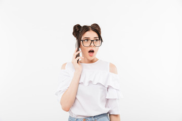 Image of shocked or surprised woman with two buns hairstyle and dental braces wearing eyeglasses talking on cell phone with open mouth, isolated over white wall