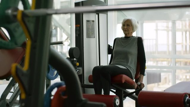 PAN of elderly woman in fitness clothes doing exercise on leg curl machine in gym