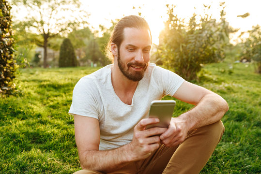 Image of caucasian handsome man in casual wear sitting on grass in green park, and using smartphone