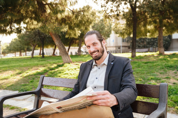 Fototapeta premium Portrait of adult handsome man in businesslike suit smiling to you, while sitting on bench in city park and reading newspaper
