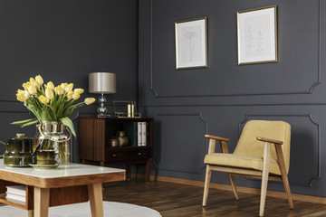 Wooden cabinet in the corner of a dark living room interior with wainscoting on the wall with posters next to a yellow armchair and table with tulips. Real photo