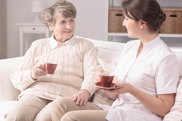 Elderly woman and her private carer sitting together on a sofa, drinking tea at home