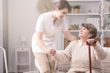 Young nurse supporting senior disabled woman with walking stick in a retirement home - obrazy, fototapety, plakaty