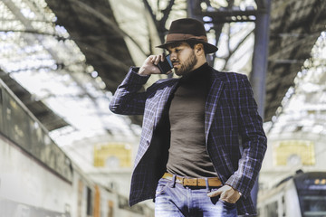 Attractive elegant young hispanic man holding smart phone in his hand and calling while he standing at the railway platform waiting for the train.