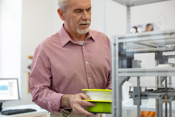 Working diligently. Pleasant senior man bringing filament for 3D printer while working in the office and carrying out printer maintenance
