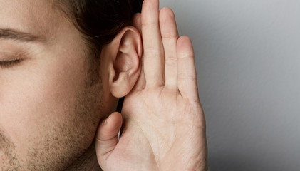 Listening male holds his hand near his ear over grey background - obrazy, fototapety, plakaty