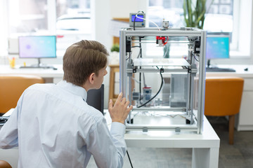 Attentive to details. Charming young office worker looking at the 3D printer and paying attention to its work algorithm