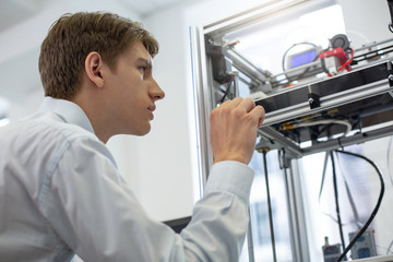 Observing all details. Pleasant young man touching the glass wall of a 3D printer while observing the printing process