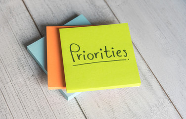 Priorities reminder handwriting on sticky note on white background, top view.