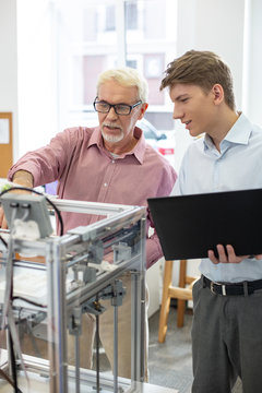 Best collaboration. Charming senior man changing 3D printer configurations together with his young intern while working in the office together