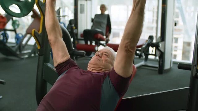 Tilt down of fit senior man with grey hair doing bench press with barbell in gym; unrecognizable elderly woman on leg curl machine in background