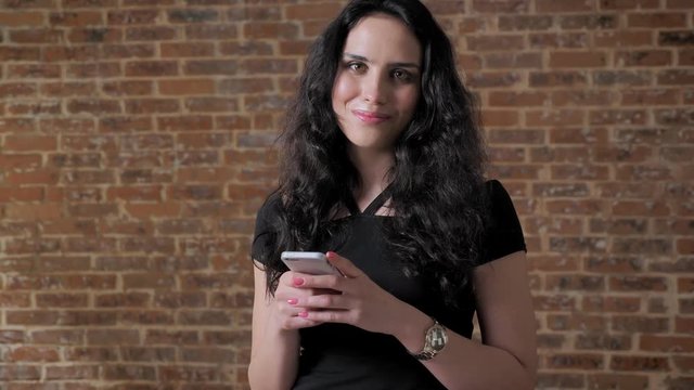 Young beautiful brunette girl surfs her smartphone, watching at camera, brick backgroung