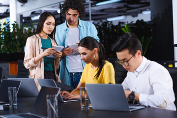 multicultural business colleagues using laptops at table while their colleagues standing with textbooks behind at modern office