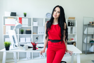 Beautiful young girl in a red suit is standing in the office, leaning on the table.