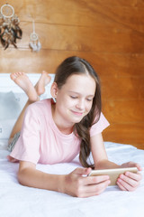 Cute preteen girl in pink t-shirt and shorts lying on bed with cell phone in boho style room against wooden wall and dreamcatchers. Scandinavian interior, modern lifestyle concept. Text space