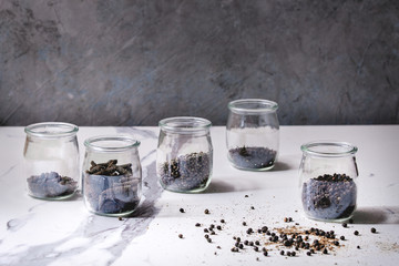 Variety of different black peppers allspice, pimento, long pepper, monks pepper, peppercorns and ground powder in glass jars on white marble kitchen table.