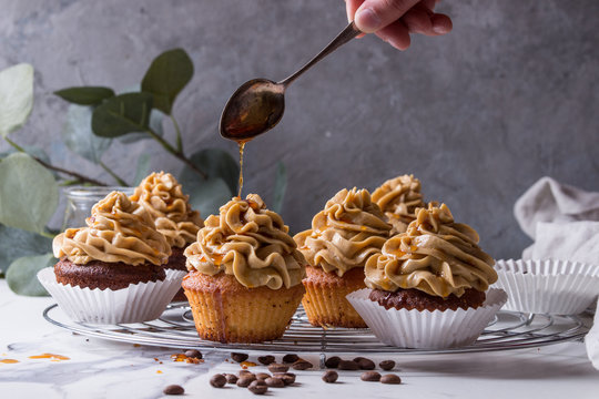 Fresh baked homemade cupcakes with coffee buttercream and pouring from spoon caramel standing on cooling rack with eucalyptus branch and coffee beans above over white marble kitchen table.