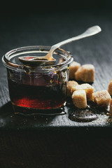 Homemade liquid transparent brown sugar caramel in glass jar standing on black wooden board with spoon and can sugar cubes. Close up. Day light